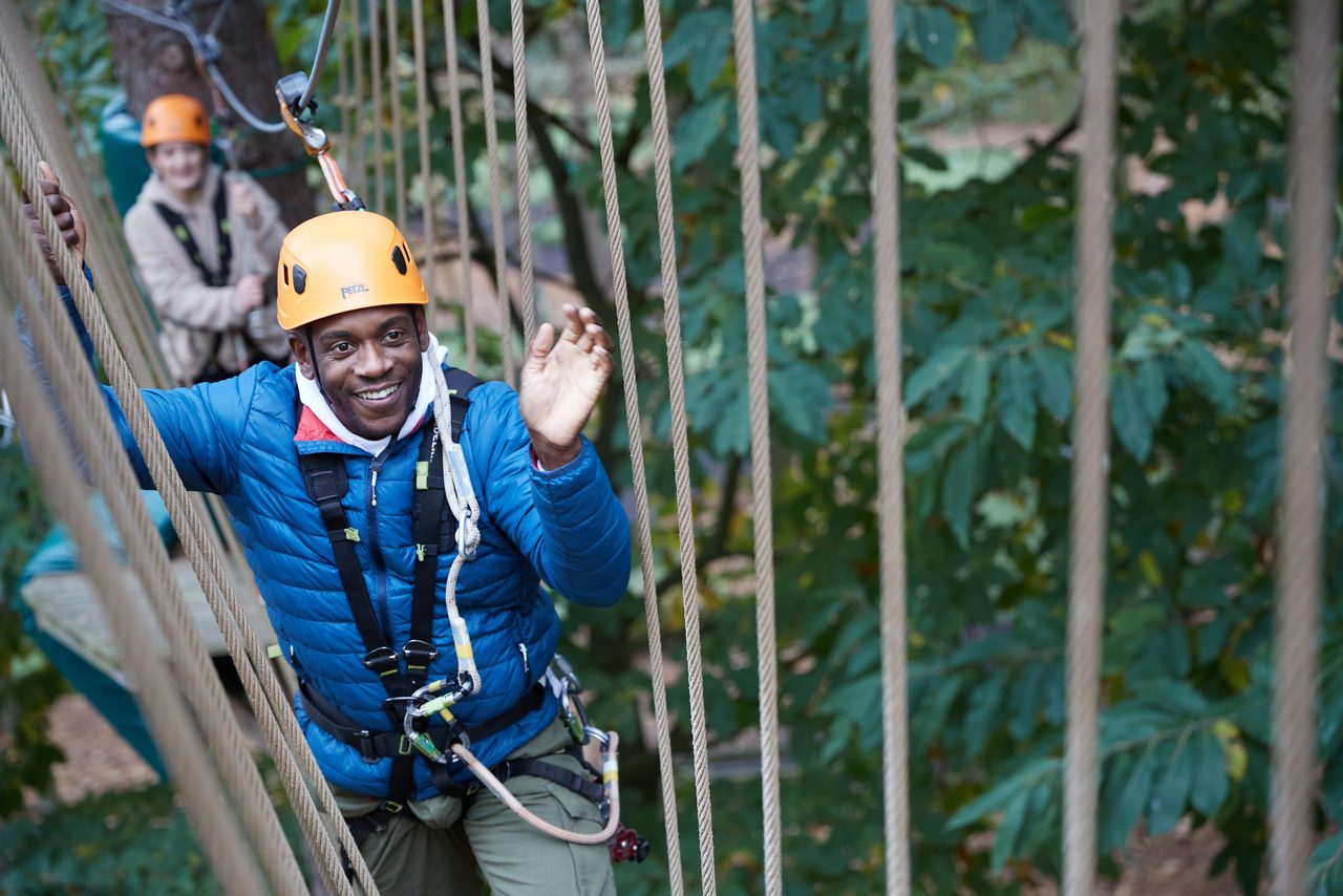 Person in a yellow helmet and safety harness crosses a suspended rope bridge, gripping ropes for balance, within a treetop adventure course among tall pine trees in warm sunlight.